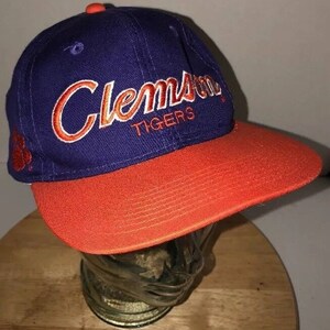 Puede incluir: Gorra de béisbol con visera naranja curvada y corona azul. La palabra "Clemson" está bordada en letras naranjas en la parte delantera, con "TIGERS" debajo. Una huella de pata naranja está en el lateral.