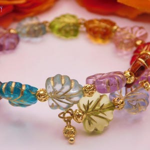 May include: A colorful beaded bracelet with various shaped beads in shades of red, pink, blue, green, and purple. The beads are connected with gold-colored accents. The bracelet is displayed on a light pink surface.