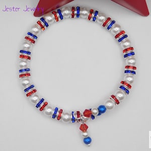 May include: A wrap bracelet featuring alternating white faux pearls and silver-toned spacers set with red and blue crystals. The bracelet includes a dangling charm composed of a red crystal bead, a white pearl, and a blue bead. Text reads Mad Jester Jewelry.