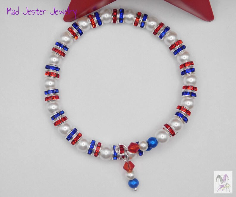 May include: A wrap bracelet featuring alternating white faux pearls and silver-toned spacers set with red and blue crystals. The bracelet includes a dangling charm composed of a red crystal bead, a white pearl, and a blue bead. Text reads Mad Jester Jewelry.