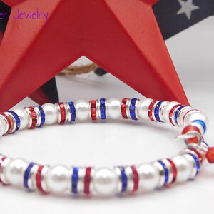May include: A stretch bracelet featuring alternating white faux pearls and silver-tone spacers set with red and blue crystals. The bracelet includes a small dangling charm with a red crystal and a blue faux pearl. The text Mad Jester Jewelry is visible.
