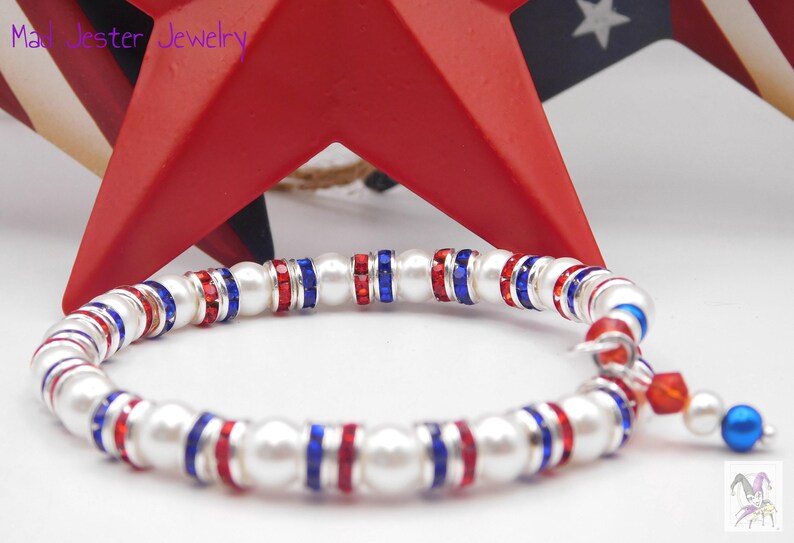 May include: A stretch bracelet featuring alternating white faux pearls and silver-tone spacers set with red and blue crystals. The bracelet includes a small dangling charm with a red crystal and a blue faux pearl. The text Mad Jester Jewelry is visible.
