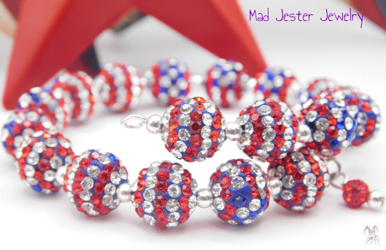 May include: A spiral bracelet featuring spherical beads encrusted with red, white, and blue rhinestones, separated by small silver-toned metal spacers. The text Mad Jester Jewelry is visible in the upper right corner of the image.