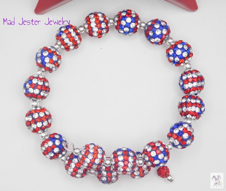 May include: A wrap bracelet featuring spherical beads covered in red, white, and blue crystals. The beads are arranged in a circular pattern with silver-toned spacers. The text Mad Jester Jewelry is visible in the upper left corner of the image.