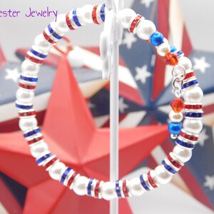 May include: A wrap bracelet featuring white faux pearls alternating with silver-tone spacers containing red and blue crystals. The bracelet includes a dangling charm with red and blue beads. The text Mad Jester Jewelry appears in the upper left corner.