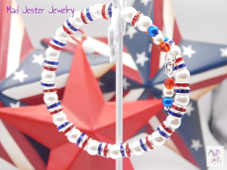 May include: A wrap bracelet featuring white faux pearls alternating with silver-tone spacers containing red and blue crystals. The bracelet includes a dangling charm with red and blue beads. The text Mad Jester Jewelry appears in the upper left corner.