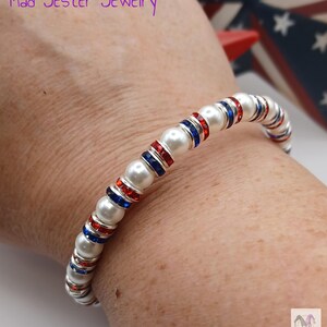 May include: A beaded bracelet featuring alternating white faux pearls and silver-toned spacers inlaid with small red and blue crystals. The text Mad Jester Jewelry is visible in the upper left corner, and a small jester icon is in the bottom right.