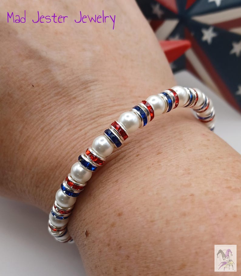 May include: A beaded bracelet featuring alternating white faux pearls and silver-toned spacers inlaid with small red and blue crystals. The text Mad Jester Jewelry is visible in the upper left corner, and a small jester icon is in the bottom right.