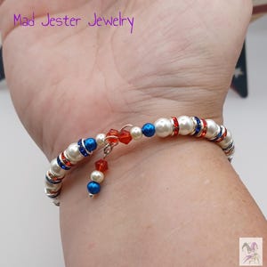 May include: A wrap bracelet featuring white faux pearls, metallic silver spacers, and alternating red and blue crystal beads. The design includes a dangling charm with a red crystal and a blue bead. The text Mad Jester Jewelry is visible.