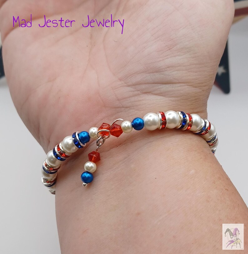 May include: A wrap bracelet featuring white faux pearls, metallic silver spacers, and alternating red and blue crystal beads. The design includes a dangling charm with a red crystal and a blue bead. The text Mad Jester Jewelry is visible.