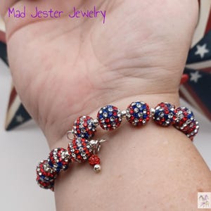 May include: A beaded bracelet featuring spherical beads encrusted with red, white, and blue rhinestones. The bracelet includes a small dangling charm with a single matching bead and a silver-toned metal clasp. The text Mad Jester Jewelry is visible.