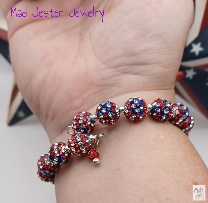 May include: A beaded bracelet featuring spherical beads encrusted with red, white, and blue rhinestones. The bracelet includes a small dangling charm with a single matching bead and a silver-toned metal clasp. The text Mad Jester Jewelry is visible.