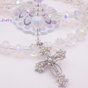 May include: A white crystal beaded necklace with a silver cross pendant adorned with small white pearls. The cross is detailed with intricate designs and has a delicate, feminine feel.