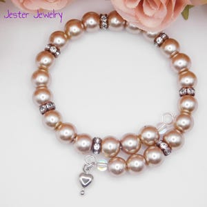 May include: A wrap bracelet featuring light brown, lustrous beads interspersed with small, clear rhinestone-encrusted spacers. A small, silver-toned heart charm hangs from one end of the wire coil. The text Mad Jester Jewelry is visible.
