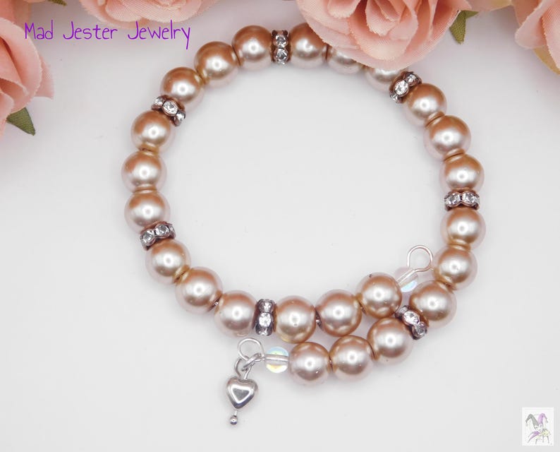 May include: A wrap bracelet featuring light brown, lustrous beads interspersed with small, clear rhinestone-encrusted spacers. A small, silver-toned heart charm hangs from one end of the wire coil. The text Mad Jester Jewelry is visible.