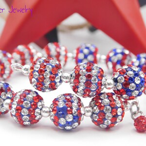 May include: A wrap bracelet featuring spherical beads encrusted with red, white, and blue rhinestones, separated by small silver-toned metal spacers. The text Mad Jester Jewelry is visible in the upper left corner of the image.