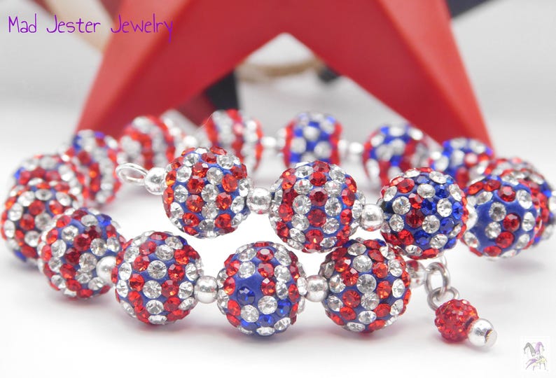May include: A wrap bracelet featuring spherical beads encrusted with red, white, and blue rhinestones, separated by small silver-toned metal spacers. The text Mad Jester Jewelry is visible in the upper left corner of the image.