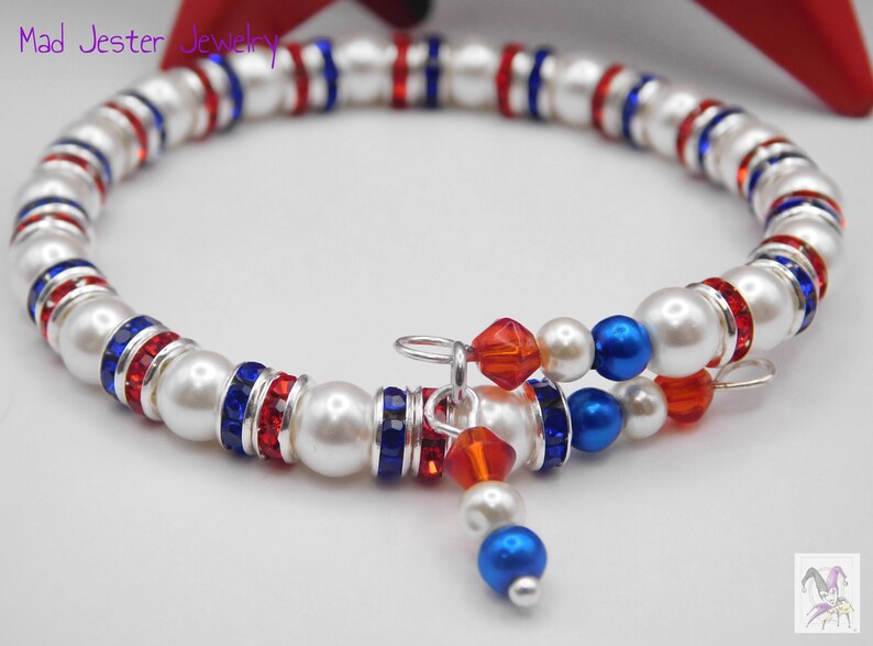 May include: A wrap bracelet featuring white faux pearls, red and blue crystal rondelles, and silver-tone spacers. The design includes dangling charms with red and blue beads. The text Mad Jester Jewelry is visible in the upper left corner.