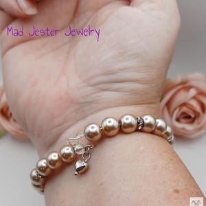 May include: A wrap bracelet featuring champagne-colored round beads, interspersed with small silver-toned spacers and a single dangling silver-toned heart charm. The text Mad Jester Jewelry is visible in purple font above the bracelet.