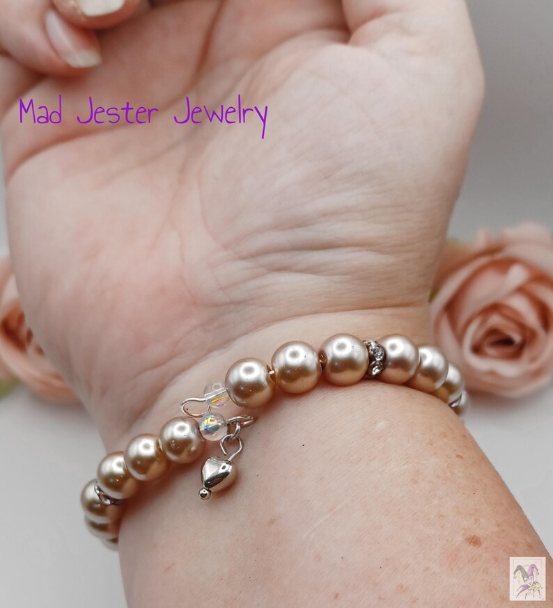 May include: A wrap bracelet featuring champagne-colored round beads, interspersed with small silver-toned spacers and a single dangling silver-toned heart charm. The text Mad Jester Jewelry is visible in purple font above the bracelet.