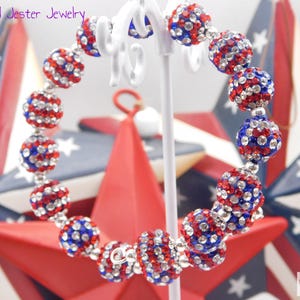 May include: A wrap bracelet featuring spherical beads encrusted with red, white, and blue rhinestones. The beads are arranged in a circular pattern on a white display stand. The text Mad Jester Jewelry is visible in the upper left corner.