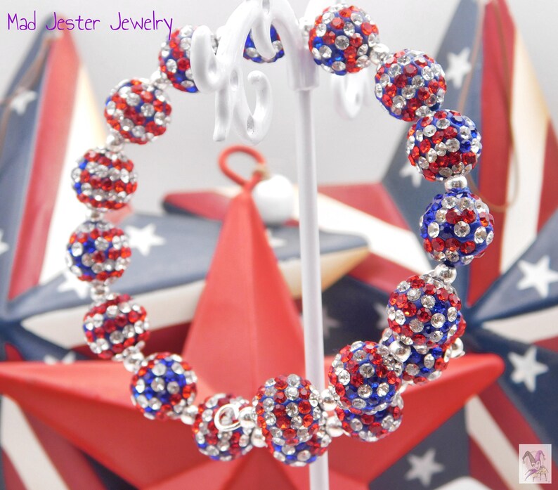 May include: A wrap bracelet featuring spherical beads encrusted with red, white, and blue rhinestones. The beads are arranged in a circular pattern on a white display stand. The text Mad Jester Jewelry is visible in the upper left corner.