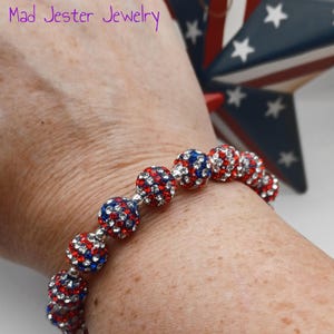 May include: A stretch bracelet featuring spherical beads covered in small red, white, and blue crystals. The text Mad Jester Jewelry is visible in the upper left corner. The beads are arranged in a single row, creating a vibrant, multi-colored pattern.