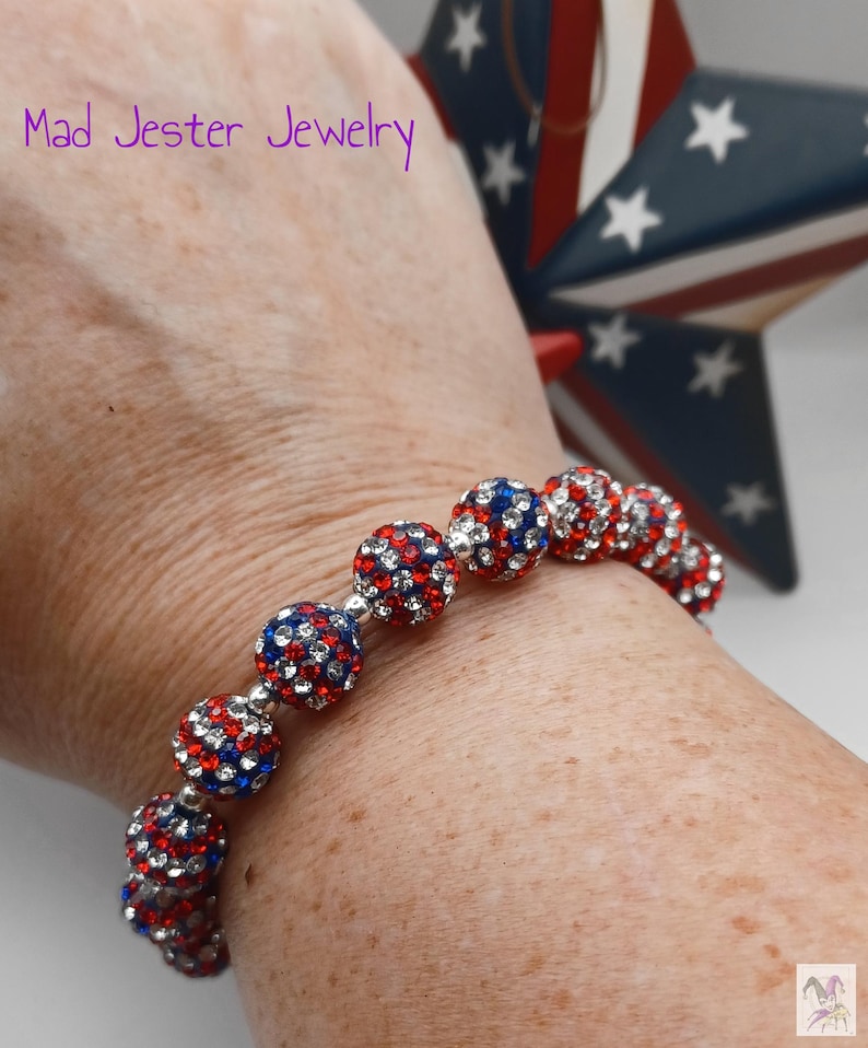 May include: A stretch bracelet featuring spherical beads covered in small red, white, and blue crystals. The text Mad Jester Jewelry is visible in the upper left corner. The beads are arranged in a single row, creating a vibrant, multi-colored pattern.