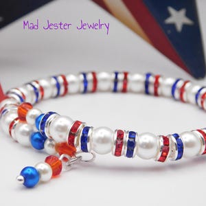 May include: A wrap bracelet featuring white faux pearls, red and blue crystal-encrusted spacers, and a dangling charm with a blue pearl, white pearl, and red crystal. The text Mad Jester Jewelry is visible in the upper portion of the image.