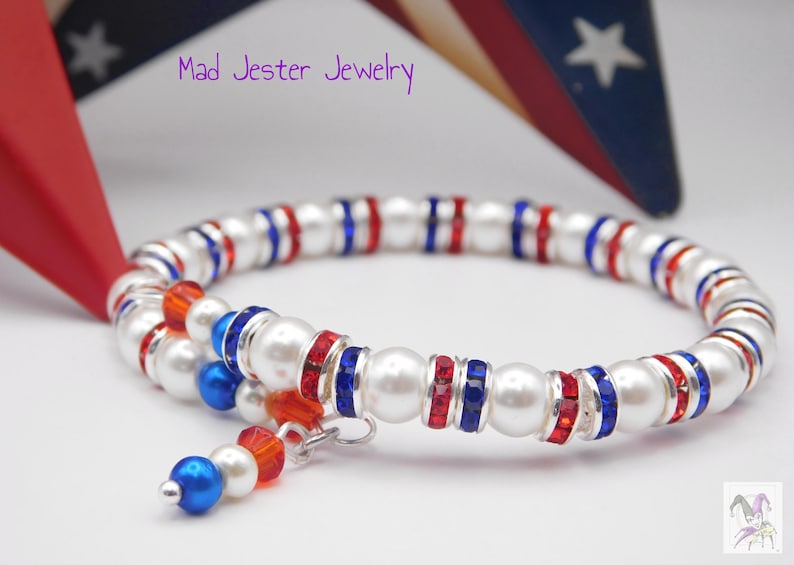 May include: A wrap bracelet featuring white faux pearls, red and blue crystal-encrusted spacers, and a dangling charm with a blue pearl, white pearl, and red crystal. The text Mad Jester Jewelry is visible in the upper portion of the image.