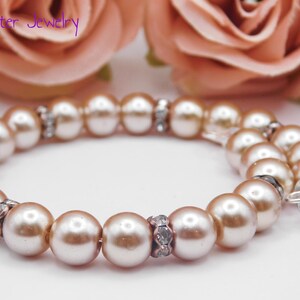 May include: A bracelet featuring round, light peach-colored pearls interspersed with small, silver-toned spacers encrusted with clear crystals. A small, silver-toned heart-shaped charm hangs from one end. The text Mad Jester Jewelry is visible.