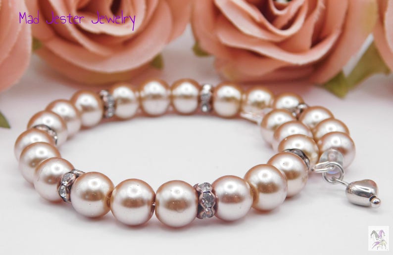 May include: A bracelet featuring round, light peach-colored pearls interspersed with small, silver-toned spacers encrusted with clear crystals. A small, silver-toned heart-shaped charm hangs from one end. The text Mad Jester Jewelry is visible.