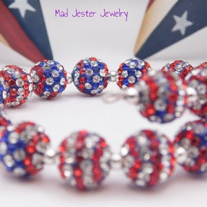 May include: A spiral bracelet featuring round beads encrusted with red, white, and blue crystals. The beads are separated by small silver-toned spacers. The text Mad Jester Jewelry is visible at the top, and a small jester icon is in the corner.
