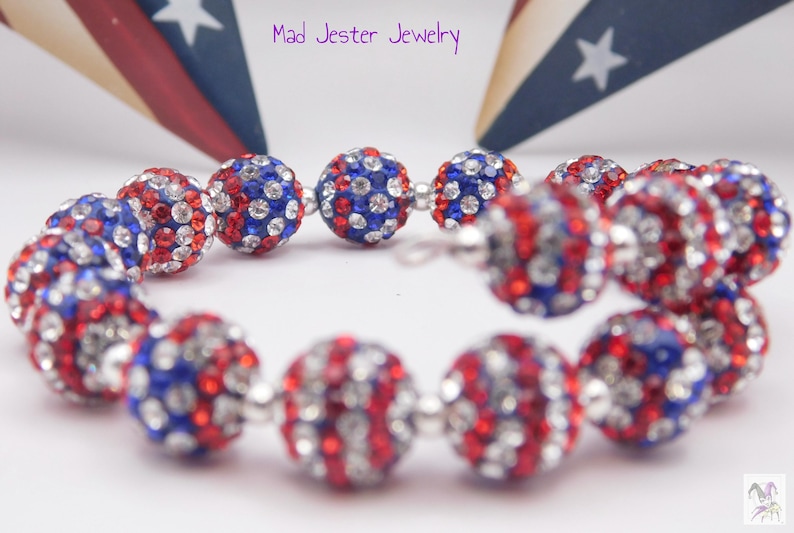 May include: A spiral bracelet featuring round beads encrusted with red, white, and blue crystals. The beads are separated by small silver-toned spacers. The text Mad Jester Jewelry is visible at the top, and a small jester icon is in the corner.
