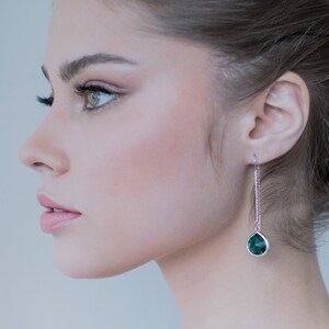 Może przedstawiać: Kobieta z włosami upiętymi w kok nosi białą sukienkę bez rękawów i szmaragdowo zielone kolczyki w kształcie łzy.