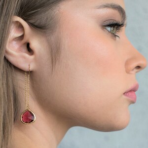 May include: A woman with long brown hair wearing a pair of gold dangle earrings with a teardrop-shaped red gemstone.
