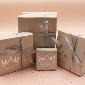 May include: Four beige gift boxes with silver ribbon tied in bows. The boxes are stacked on top of each other. The smallest box has a silver crown and the text "Princess K" on it. The other boxes have the text "Princess K" on them.