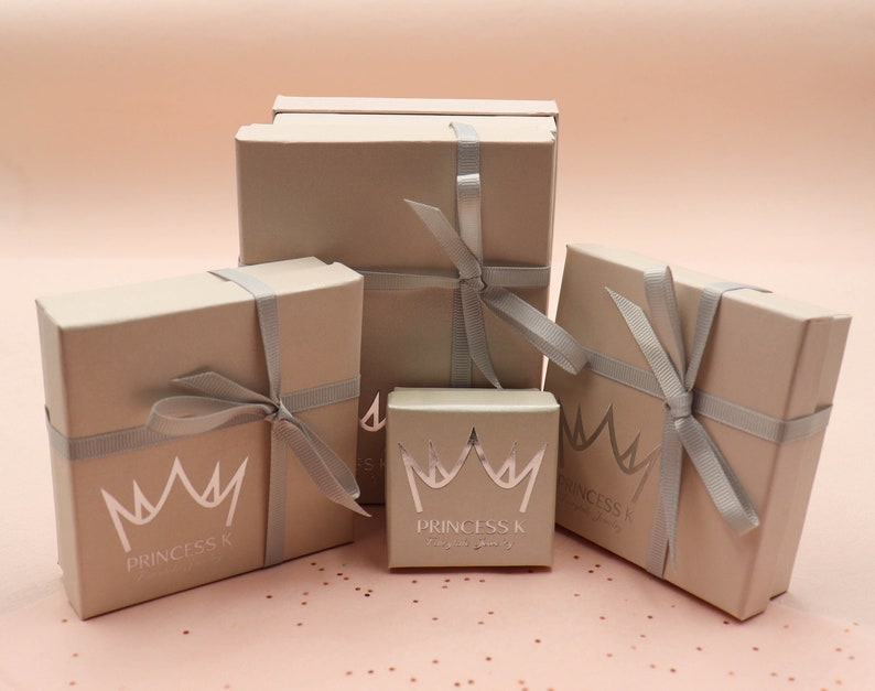 May include: Four beige gift boxes with silver ribbon tied in bows. The boxes are stacked on top of each other. The smallest box has a silver crown and the text "Princess K" on it. The other boxes have the text "Princess K" on them.