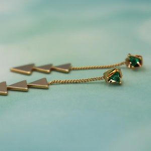 Pode incluir: Um par de brincos de ouro com uma corrente e três pingentes em forma de triângulo. Cada brinco tem uma gema verde no final da corrente.