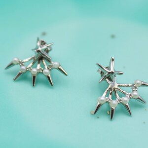 May include: A pair of silver earrings with a star design and pearl accents. The earrings are on a light blue background.