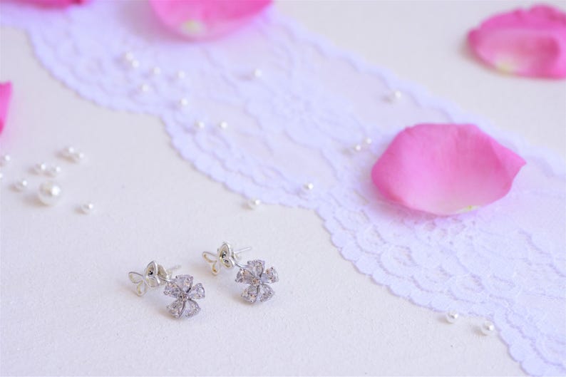 May include: A pair of silver earrings with a delicate flower design and clear sparkling stones. The earrings are on a white lace background with pink rose petals.