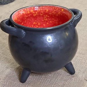 May include: A small black cauldron with three legs and orange speckled red interior. The cauldron is made of ceramic and has two handles.