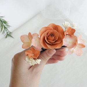 May include: A floral hair accessory featuring a large, burnt orange rose surrounded by smaller peach and white flowers. The arrangement is mounted on a black hairpin, creating a delicate and natural look. Perfect for weddings or special occasions.
