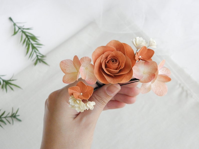 May include: A floral hair accessory featuring a large, burnt orange rose surrounded by smaller peach and white flowers. The arrangement is mounted on a black hairpin, creating a delicate and natural look. Perfect for weddings or special occasions.