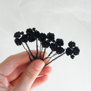 Può includere: Un gruppo di forcine per capelli floreali nere. Ogni forcina presenta un piccolo fiore scuro attaccato a una forcina in metallo nero. I fiori sembrano essere fatti di tessuto. Lo sfondo è un tessuto bianco e morbido.