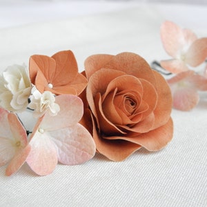 May include: A floral hair accessory featuring a large, paper-crafted rose in a warm, earthy tone, complemented by smaller blossoms in shades of peach and cream. The arrangement is delicate and suitable for weddings or special events.