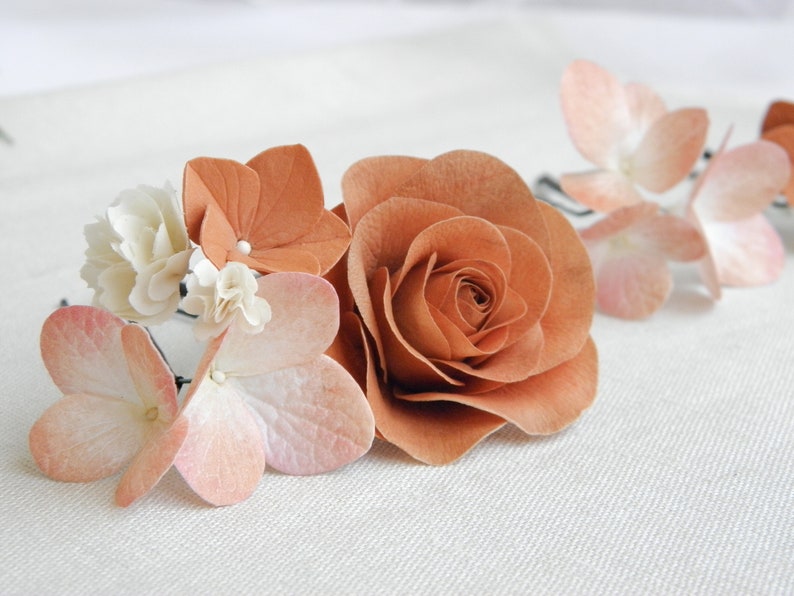 May include: A floral hair accessory featuring a large, paper-crafted rose in a warm, earthy tone, complemented by smaller blossoms in shades of peach and cream. The arrangement is delicate and suitable for weddings or special events.