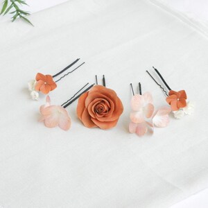 May include: A collection of hair accessories featuring floral designs. The set includes a large, burnt orange rose and several hairpins adorned with smaller orange and pink flowers. The hair accessories are arranged on a white fabric surface.