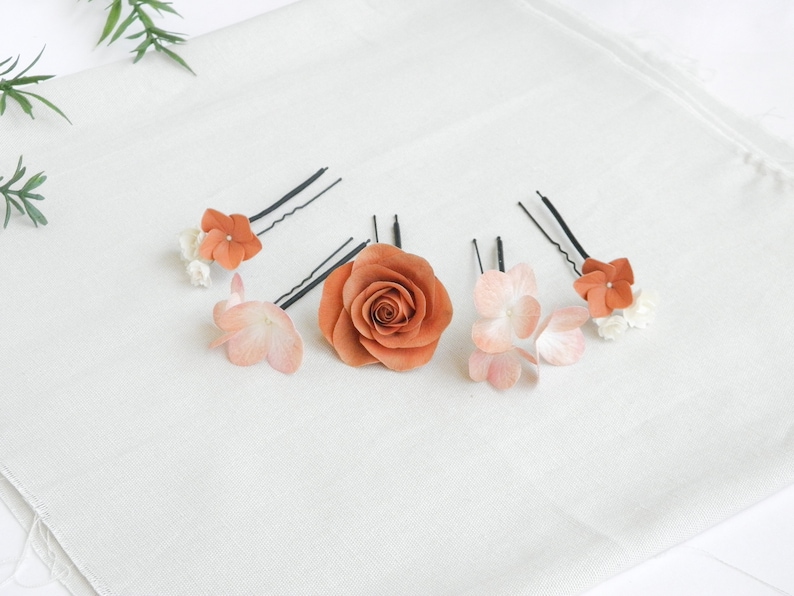 May include: A collection of hair accessories featuring floral designs. The set includes a large, burnt orange rose and several hairpins adorned with smaller orange and pink flowers. The hair accessories are arranged on a white fabric surface.