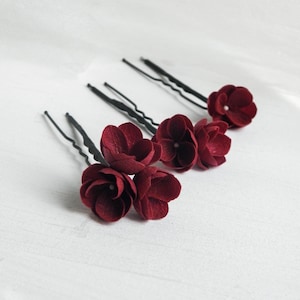 May include: Five burgundy flower hair pins. Each pin features a dark red flower with detailed petals. The pins are arranged diagonally on a white surface, showcasing the floral design and the black pin stems.